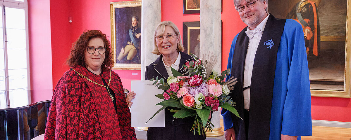 Ehrenpromotion Ursula Gather Drei Menschen stehen in einem eleganten Raum, eine Frau wurde geehrt und hält einen Blumenstrauß in der Hand.
