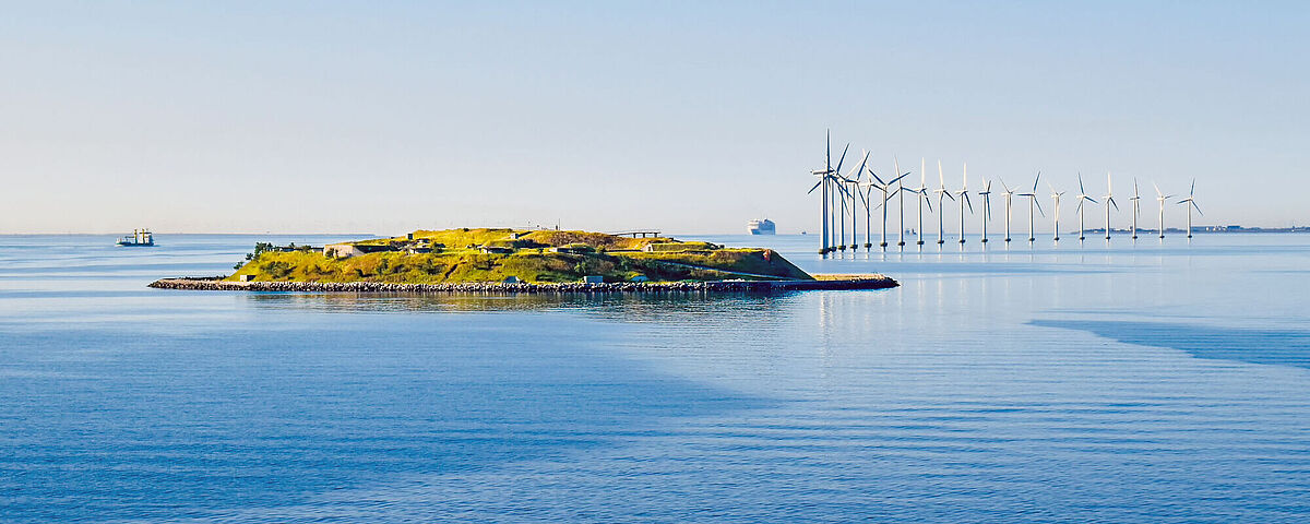 Eine Reihe Windräder steht hinter einer kleinen Insel in der Ostsee.