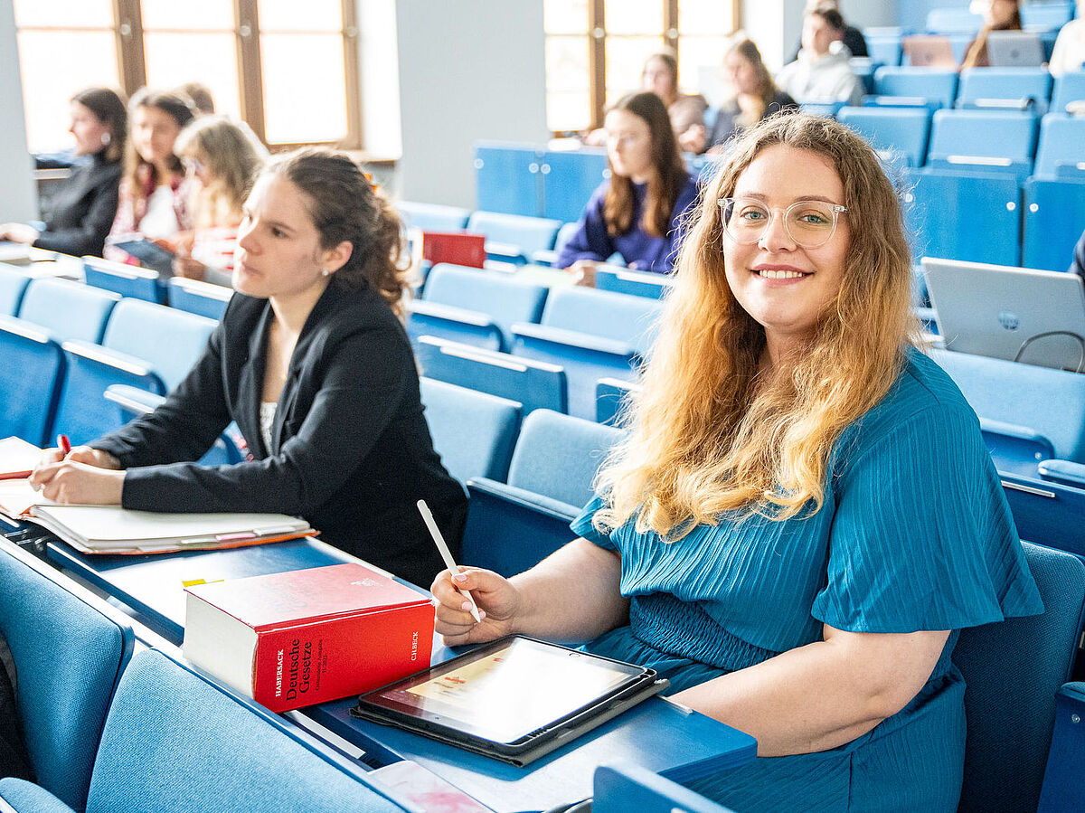 Im Hörsaal sitzen viele Studierende. Im Vordergrund sitzt eine blonde Frau in einem blauen Kleid.