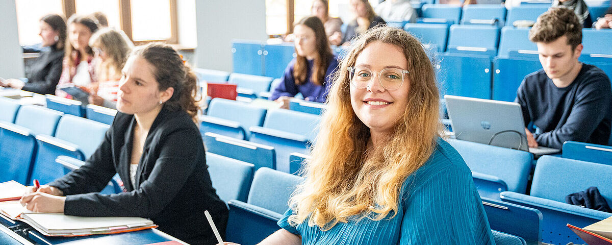 Im Hörsaal sitzen viele Studierende. Im Vordergrund sitzt eine blonde Frau in einem blauen Kleid.