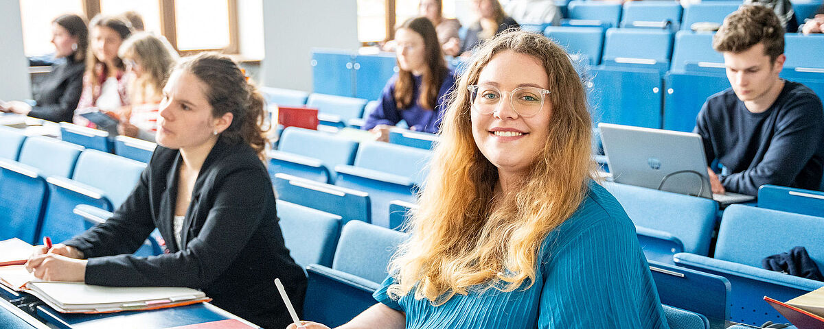 Im Hörsaal sitzen viele Studierende. Im Vordergrund sitzt eine blonde Frau in einem blauen Kleid.