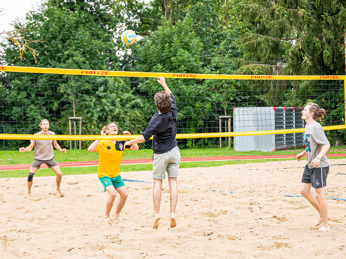 Eine Gruppe von vier jungen Männern sind sommerlich gekleidet und spielen auf einem sandigen Platz Beachvolleyball. Im Hintergrund stehen grüne Bäume.