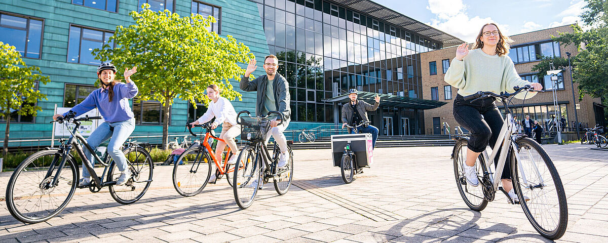 Studierende fahren mit dem Fahrrad und winken fröhlich in die Kamera. Im Hintergrund ist die Zentrale Universitätsbibliothek zu sehen.