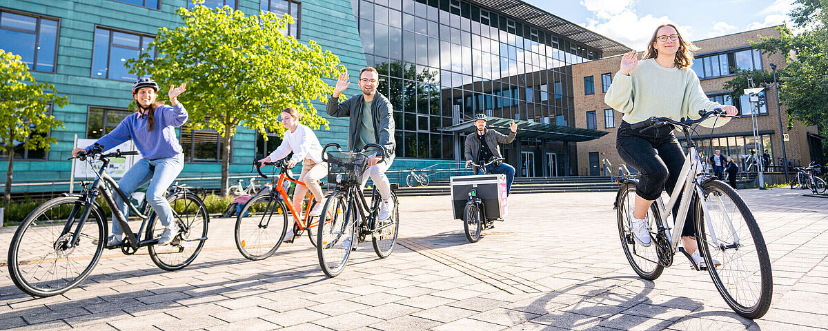 Studierende fahren mit dem Fahrrad und winken fröhlich in die Kamera. Im Hintergrund ist die Zentrale Universitätsbibliothek zu sehen.