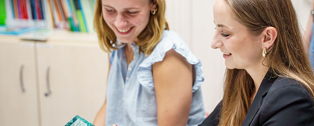 Zwei weibliche Studierende schauen sich gemeinsam ein Buch an und freuen sind dabei.